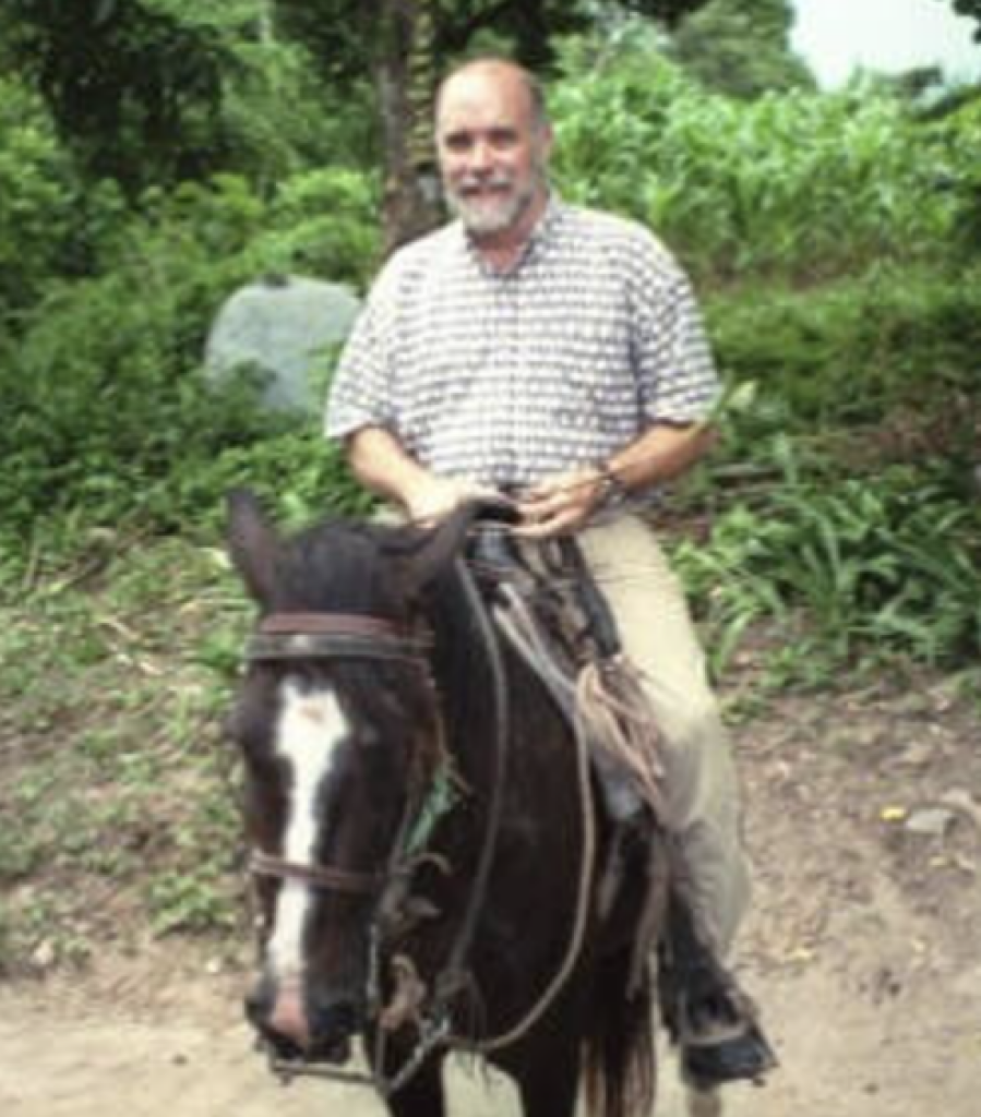 A bearded man in a checkered shirt and khaki pants sits on a horse in a lush, green outdoor setting. The background is filled with trees and plants.